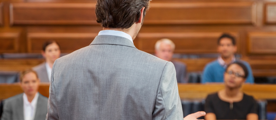  Blurred background of five (5) people sitting in a jury box in a courtroom, with a man, presumably an attorney, standing outside of the jury box facing the individuals in the jury box.