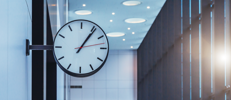 A clock hanging on a wall in a hallway with a wall of windows to the right side.